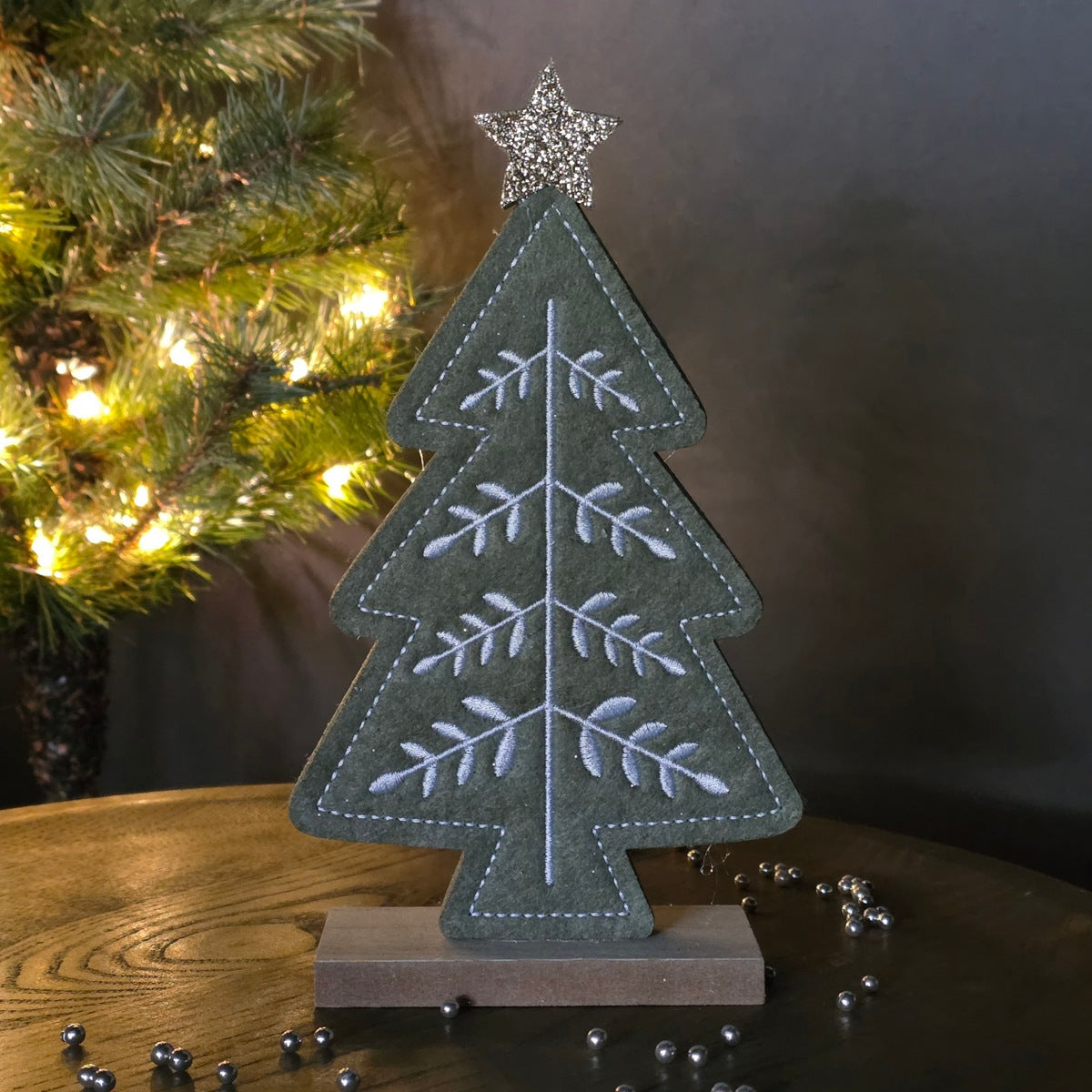 A felt Christmas tree decoration in muted green with white embroidered branches and a silver glitter star on top, displayed on a wooden base on a tabletop with warm holiday lights in the background.