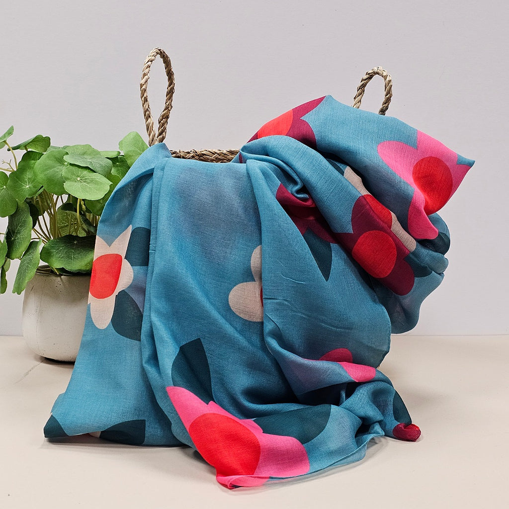 A lightweight duck egg blue scarf with large flower prints in  in deep red, pink, white, red and teal leaves, draped over a woven straw basket next to a potted green plant.
