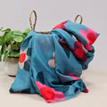 A lightweight duck egg blue scarf with large flower prints in  in deep red, pink, white, red and teal leaves, draped over a woven straw basket next to a potted green plant.