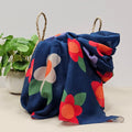 A lightweight navy scarf with large flower prints in  in red, white, purple, orange and green leaves, draped over a woven straw basket next to a potted green plant.