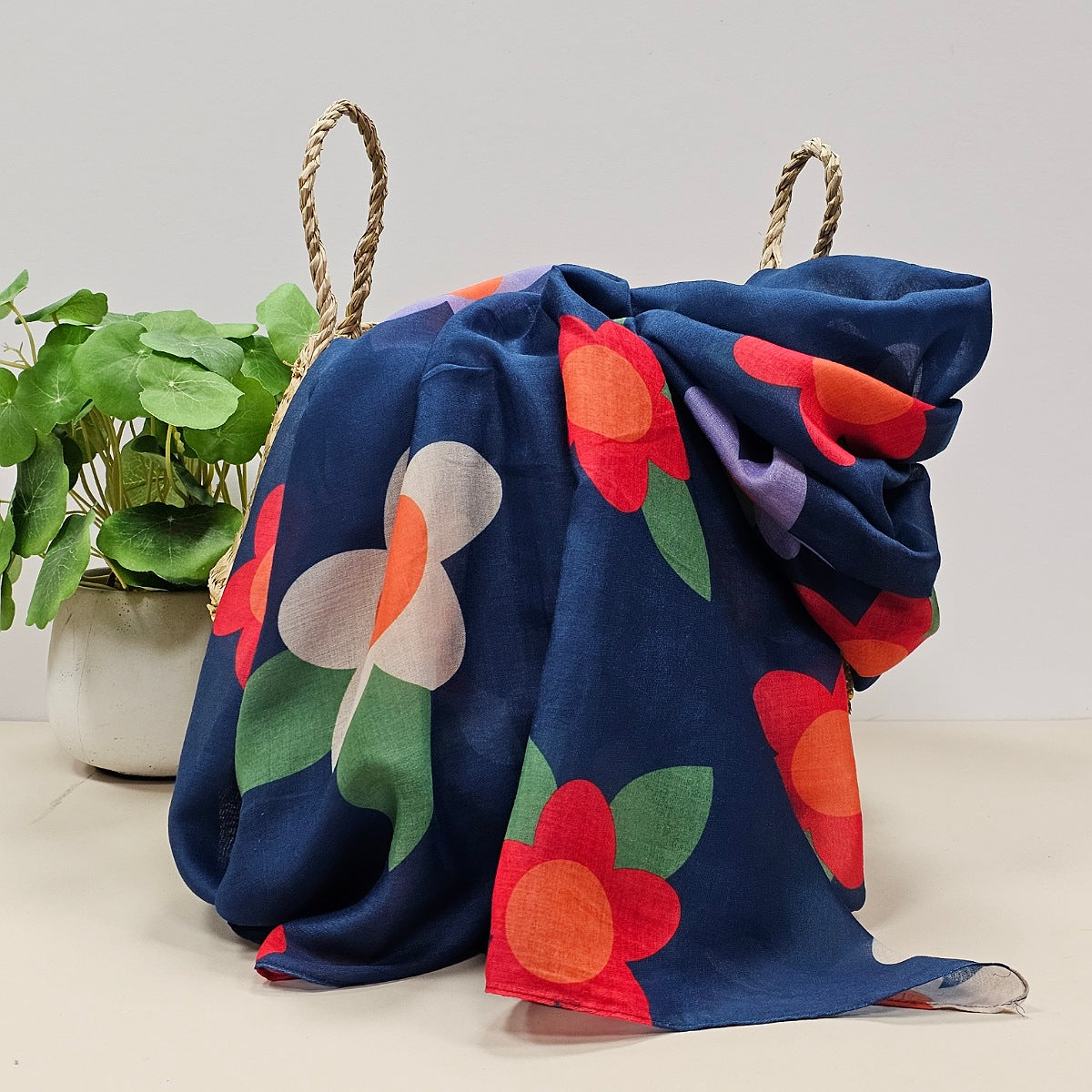 A lightweight navy scarf with large flower prints in  in red, white, purple, orange and green leaves, draped over a woven straw basket next to a potted green plant.