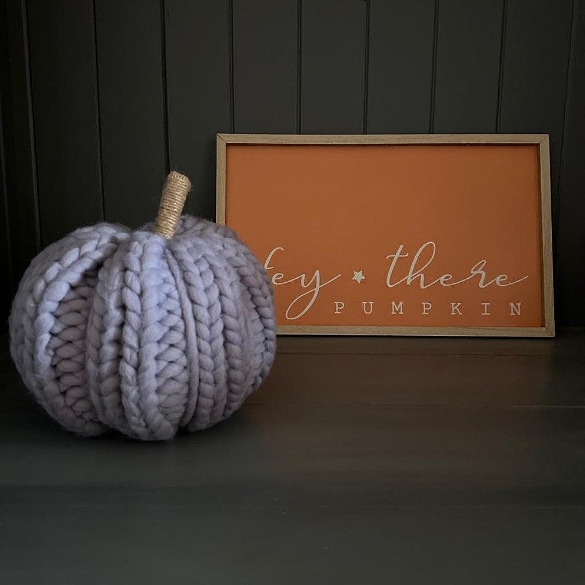 Large grey knitted wool pumpkin and string stalk next to a framed sign with 'Hey there Pumpkin' text on a dark background