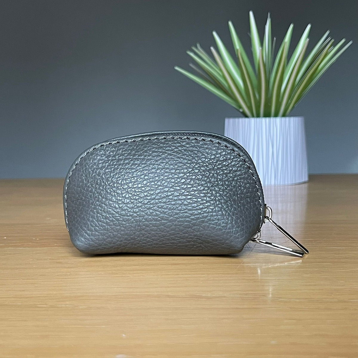 A small charcoal grey pebbled leather zip pouch with a metal key ring, resting on a light wooden table with a potted plant in the background.