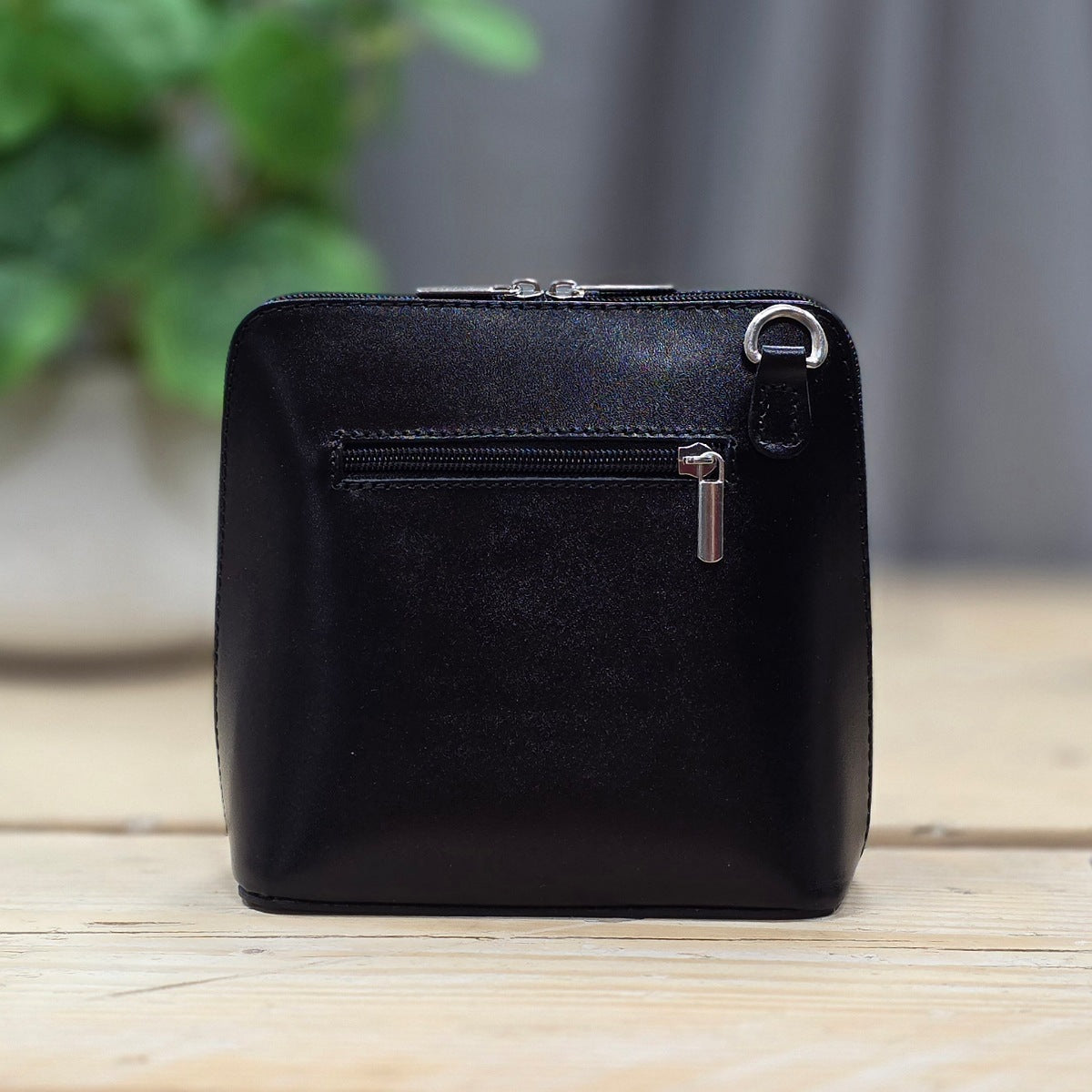 Black leather handbag with a zipper on a wooden surface with a blurred green plant in the background