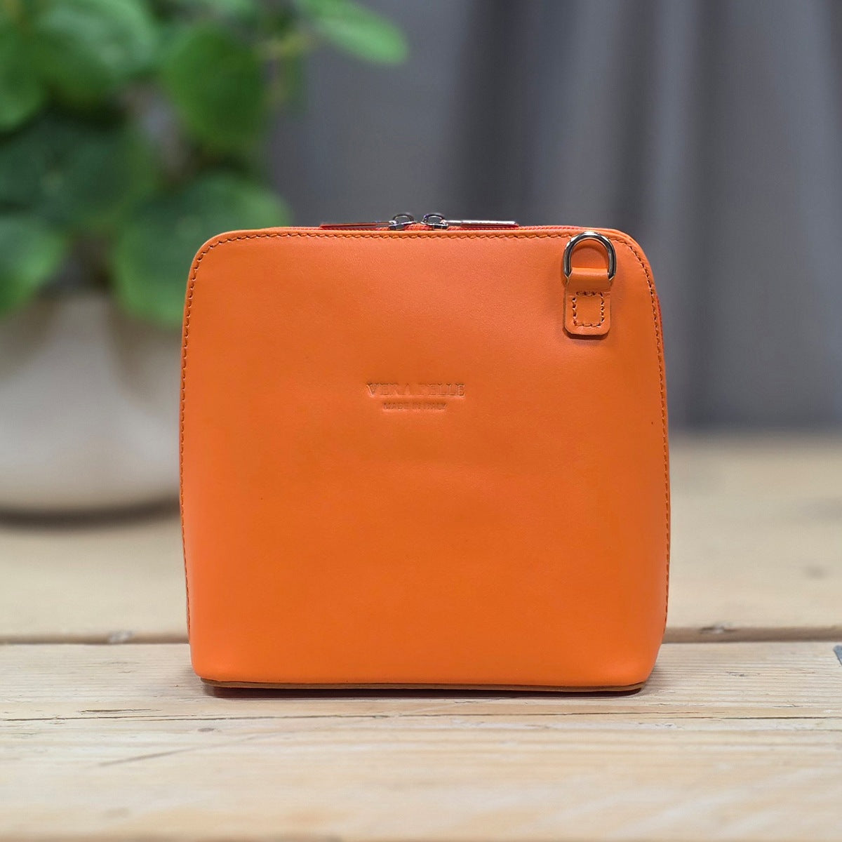 An orange leather small bag with a top zipper, metal ring detail, displayed on a wooden surface with a blurred indoor background.