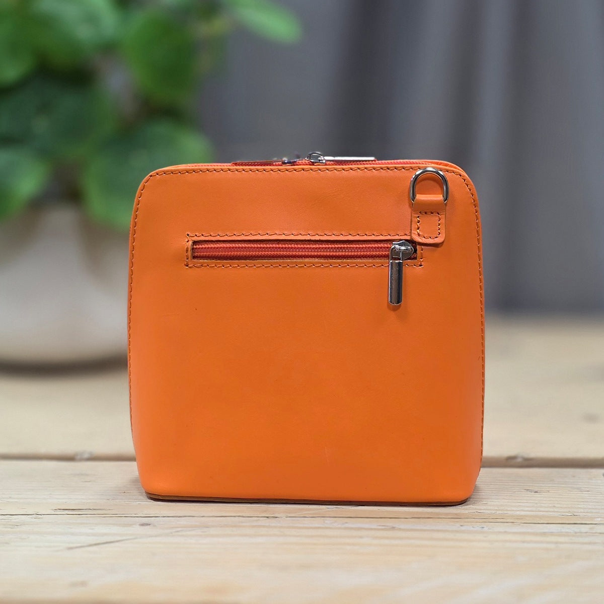 Orange leather handbag with a zipper on a wooden surface with a blurred green plant in the background