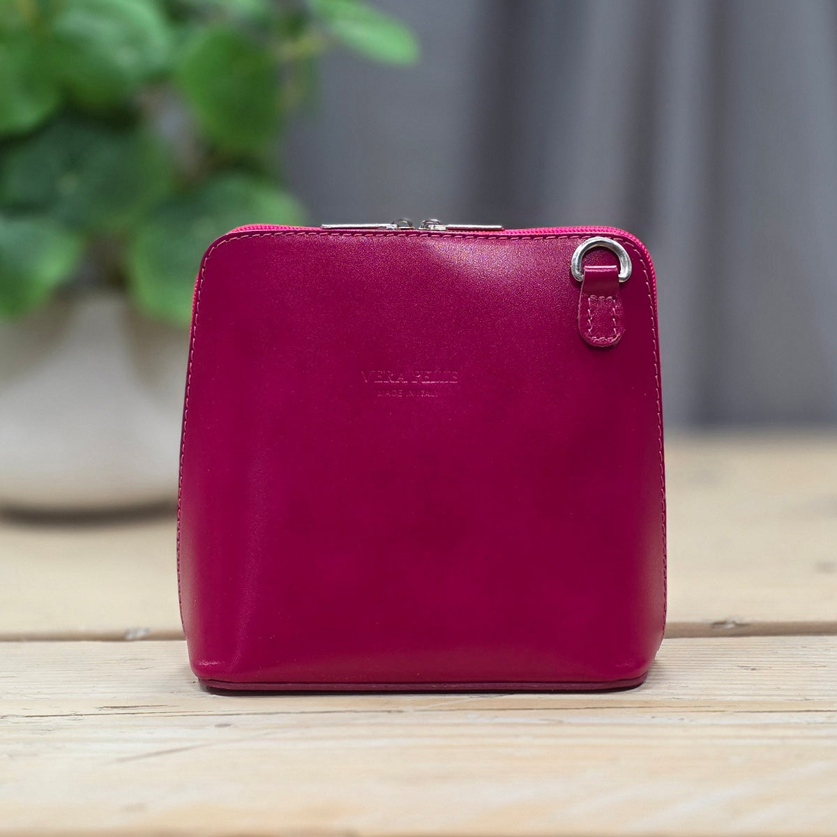 A Fuchsia pink leather zip handbag with a small silver ring detail, standing on a light wooden surface with a blurred green plant in the background.