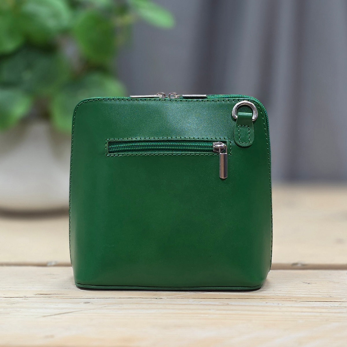 Green leather handbag with a zipper on a wooden surface with a blurred plant in the background.