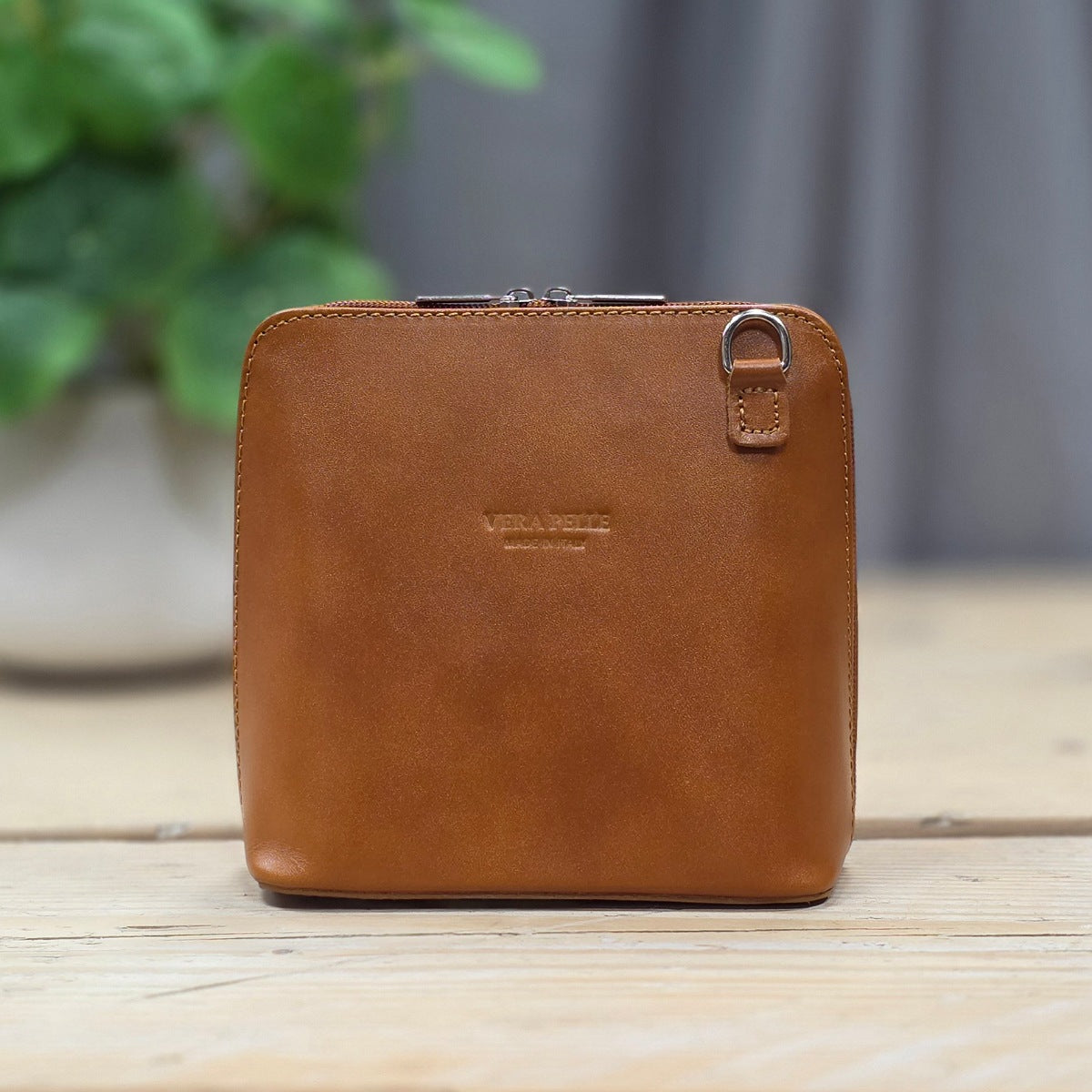 A brown leather small handbag with a front zip pocket and metal hardware, standing on a light wooden surface with a blurred plant and gray backdrop in the background.