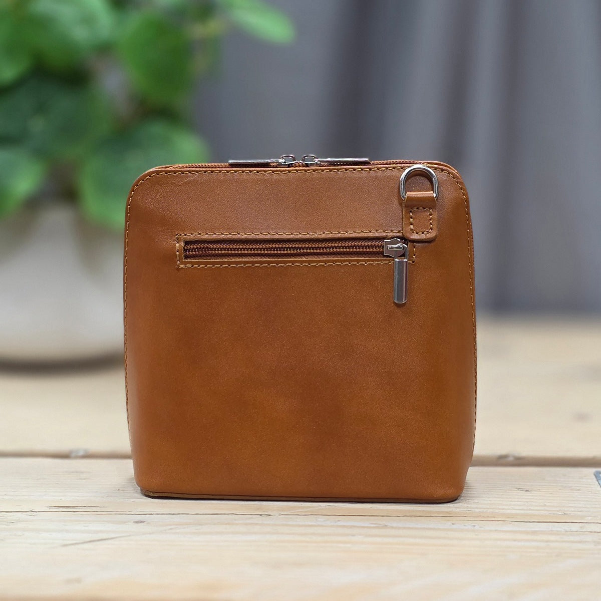 A brown leather small handbag with a front zip pocket and metal hardware, standing on a light wooden surface with a blurred plant and gray backdrop in the background.