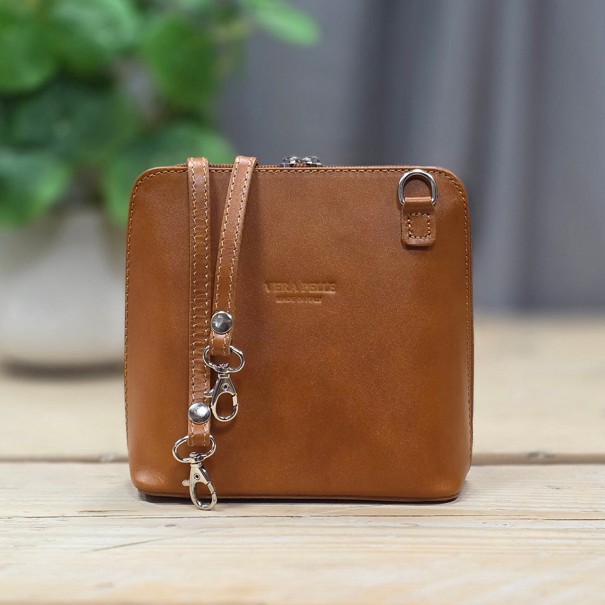 A brown leather small handbag with a front zip pocket and metal hardware, standing on a light wooden surface with a blurred plant and gray backdrop in the background.