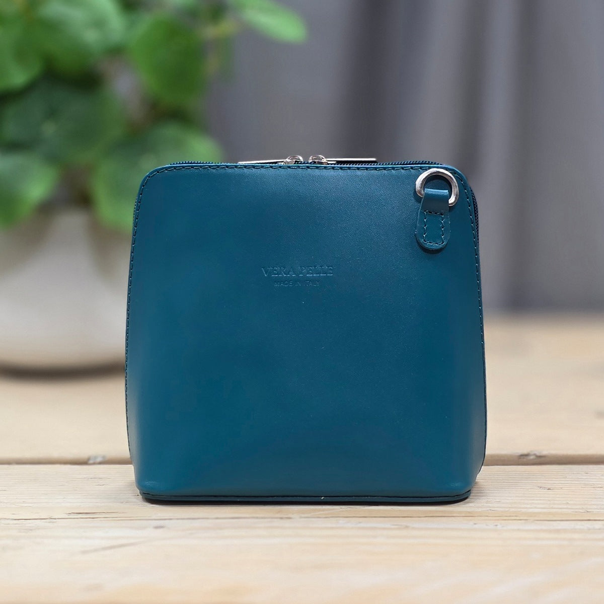 Teal handbag on a wooden surface with a blurred green plant in the background