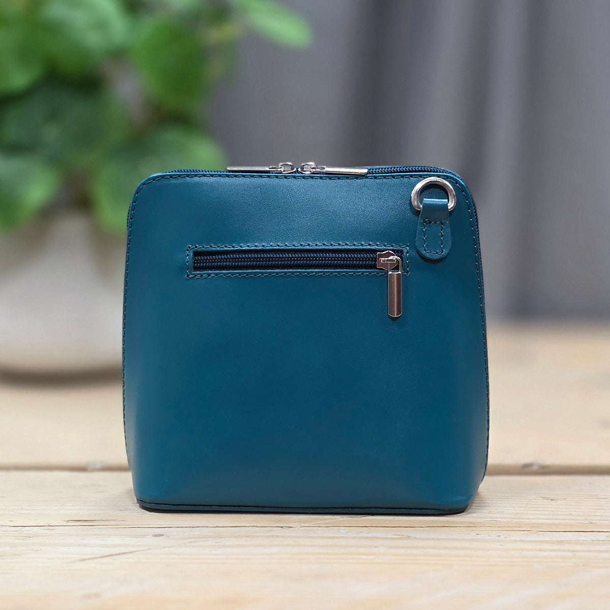 A teal blue leather zip-around wallet standing upright on a light wooden surface, with a small horizontal zip pocket and silver-tone hardware on the front. A blurred green plant and gray curtain appear in the background.