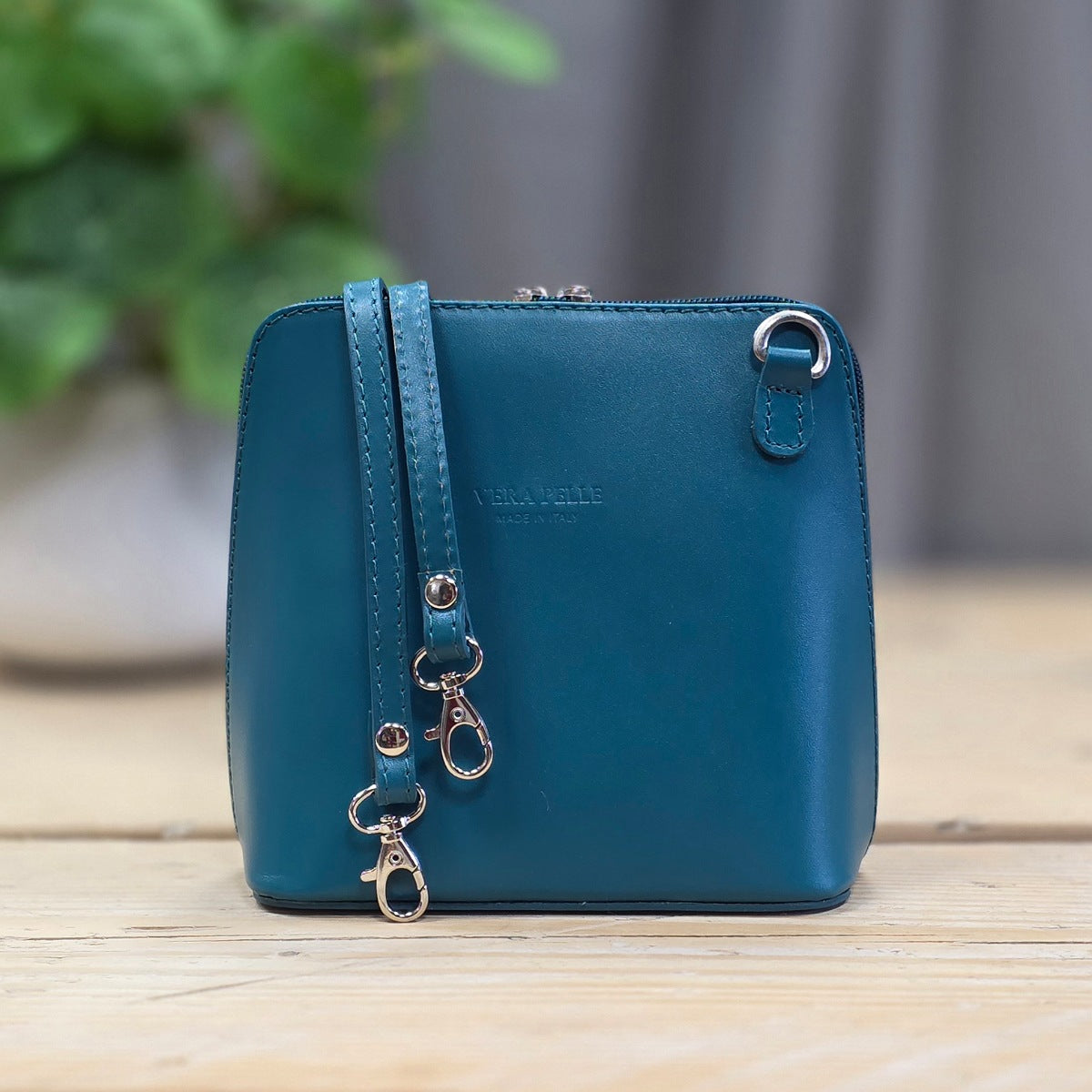 A small teal blue leather pouch with a wrist strap and silver-tone hardware, standing on a light wood surface with a blurred indoor background.