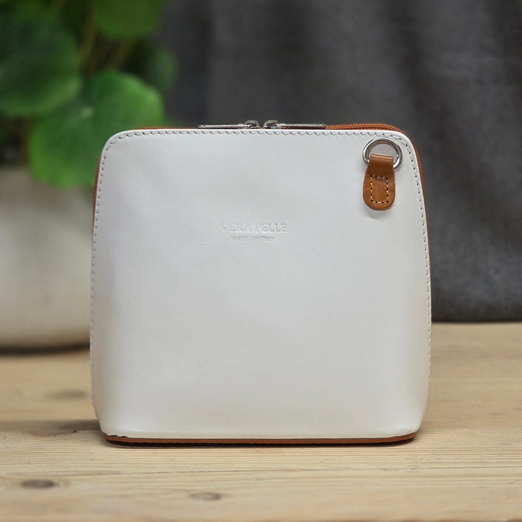 A square white leather zip pouch with a small brown leather tab and silver metal ring, standing on a light wooden surface with a blurred plant and gray fabric in the background.