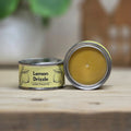 An open round metal candle tin labeled 'Lemon Drizzle' sits on a wooden surface with its lid placed beside it. The candle wax is a warm yellow-green, and the label features lemon illustrations.