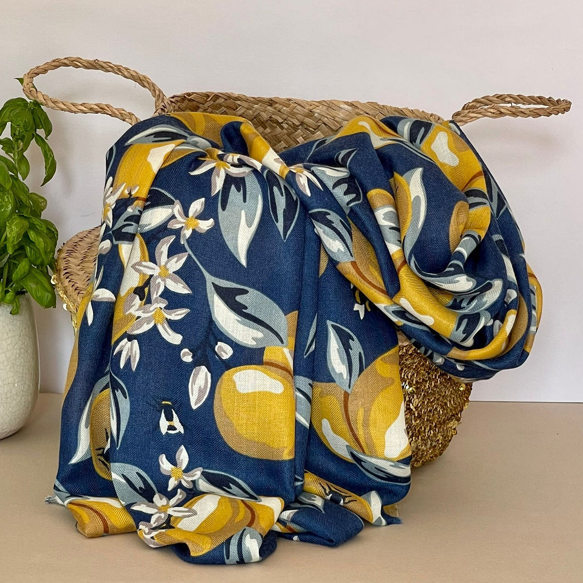 A navy blue scarf with a floral design with lemons and bees in yellow, grey, navy, white and beige, draped over a woven straw basket next to a potted green plant.
