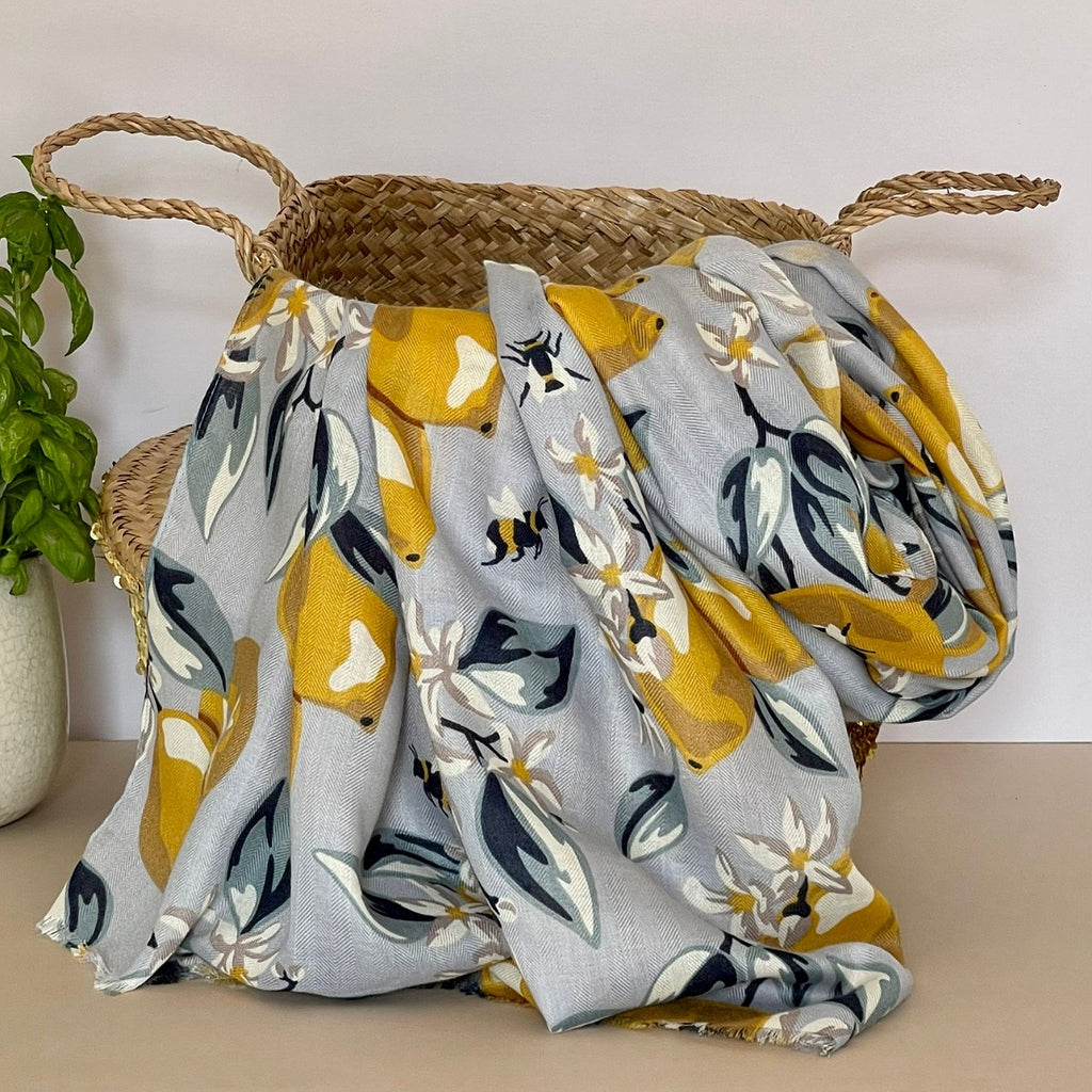 A silver scarf with a floral design with lemons and bees in yellow, navy, grey, white and beige, draped over a woven straw basket next to a potted green plant.
