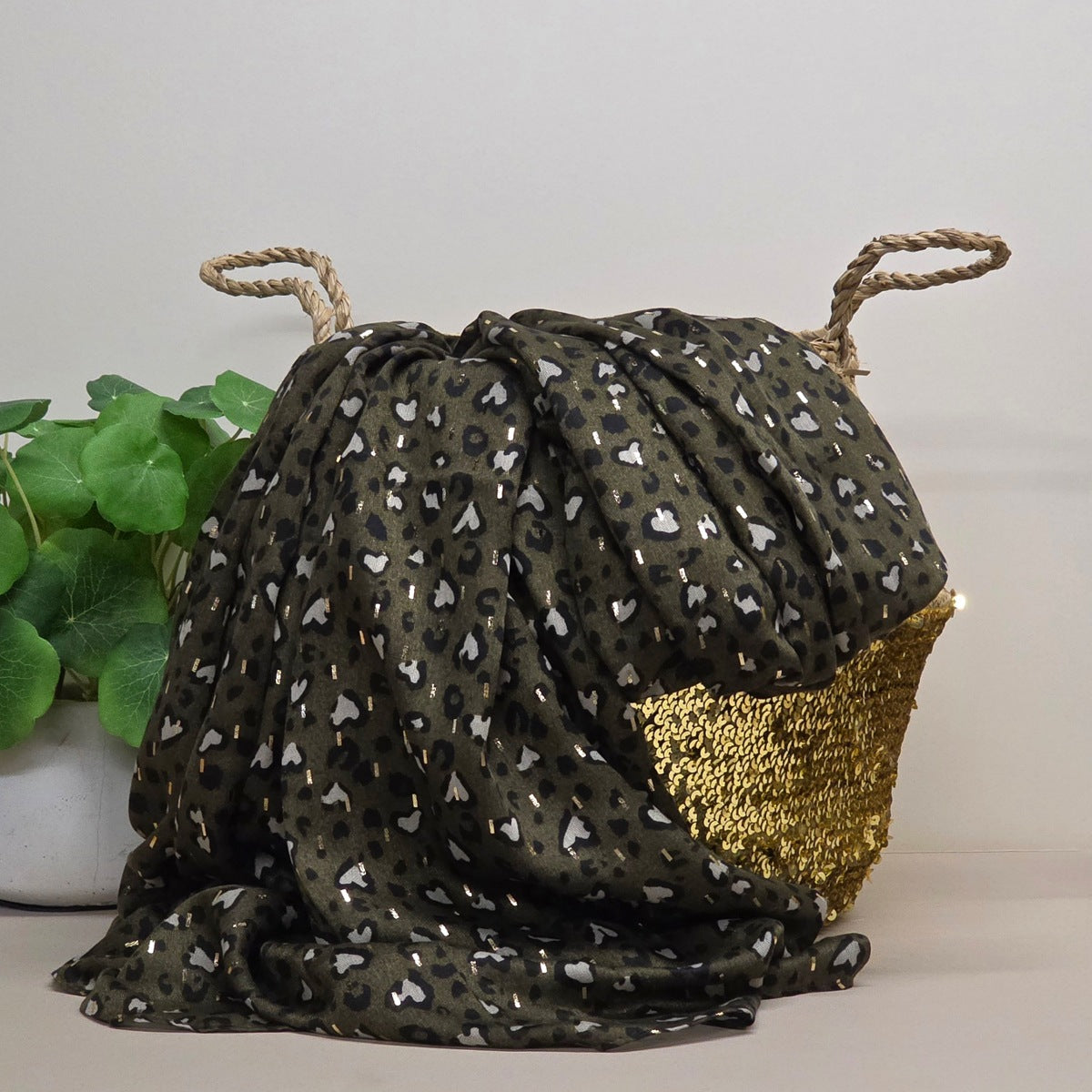 A dark olive green scarf with a leopard print pattern and a textured gold side pocket, styled next to a green potted plant against a neutral background.