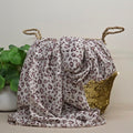 A draped leopard-print scarf or shawl in off-white with brown and muted purple spots, styled next to a woven basket with rope handles and a gold sequin accent, with a potted plant on the left.