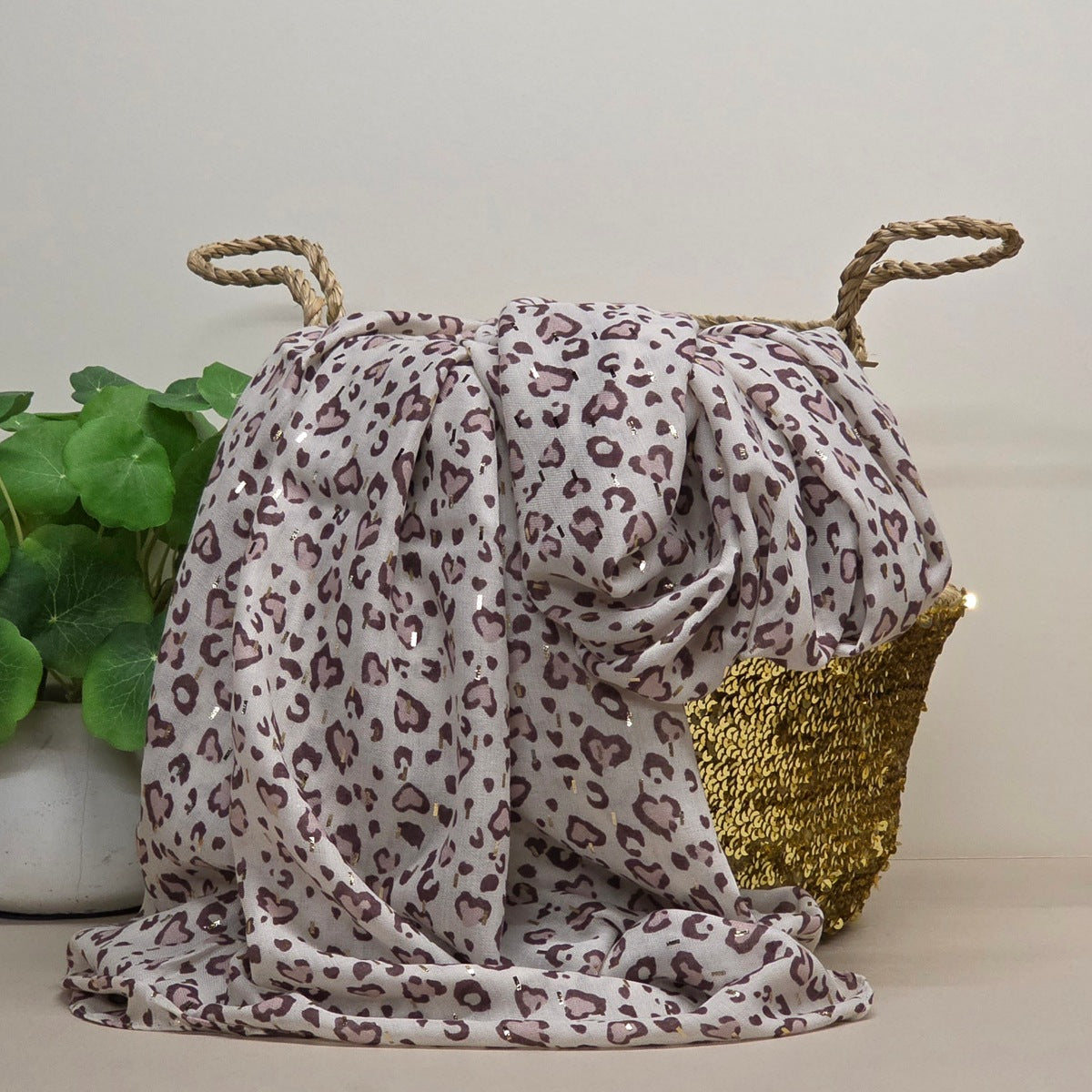 A draped leopard-print scarf or shawl in off-white with brown and muted purple spots, styled next to a woven basket with rope handles and a gold sequin accent, with a potted plant on the left.