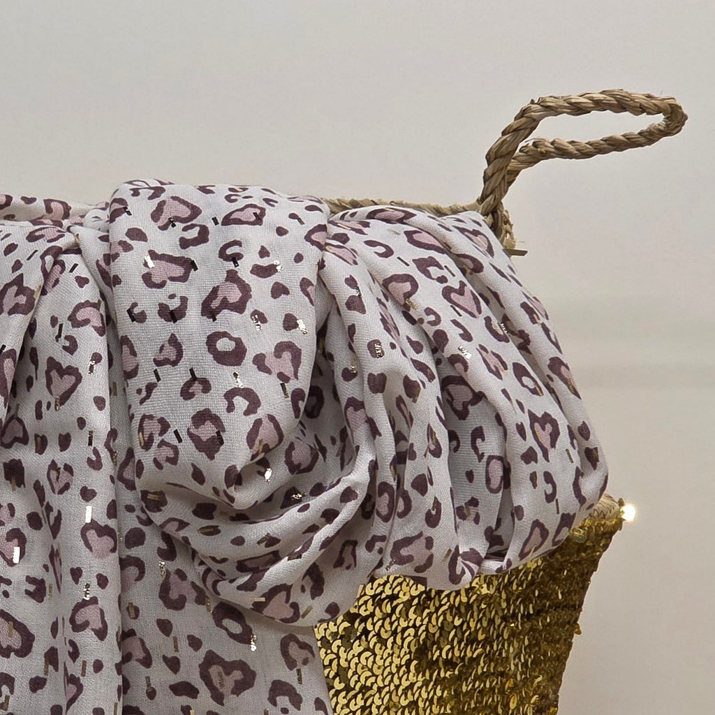 A draped leopard print fabric with a white base and dusty purple spots, styled over a gold sequined basket with a rope handle against a plain light background.