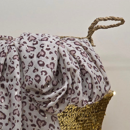 A draped leopard print fabric with a white base and dusty purple spots, styled over a gold sequined basket with a rope handle against a plain light background.