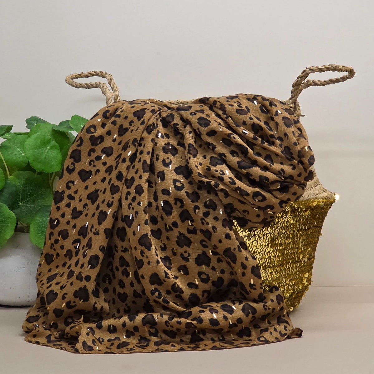 A leopard print scarf draped over a woven basket with rope handles, set against a neutral background with a green plant on the left.