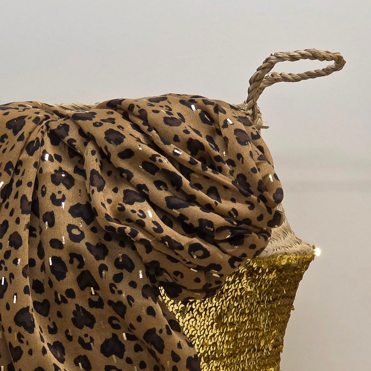 A leopard print scarf draped over a woven basket with a gold woven handle, set against a plain light grey background.