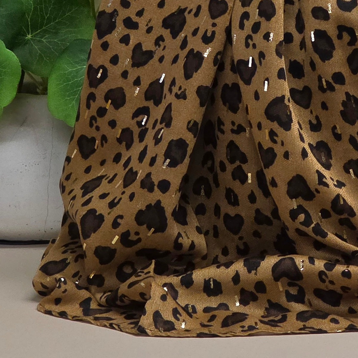 A draped leopard print fabric with a tan base, black spots, and small reflective silver accents, shown next to green leaves.