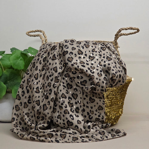 A leopard print scarf with sequins draped over a woven basket with rope handles, styled next to a green plant on a light beige surface against a neutral background.