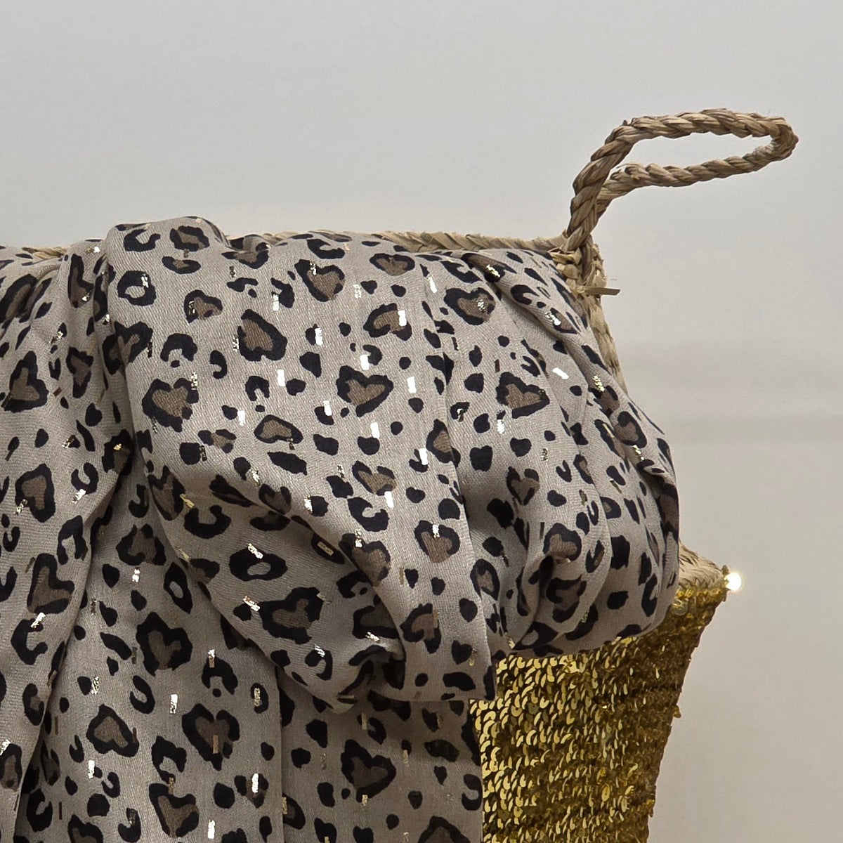 A leopard print scarf with sequins draped over a woven straw basket with a gold woven base, set against a plain light grey background.