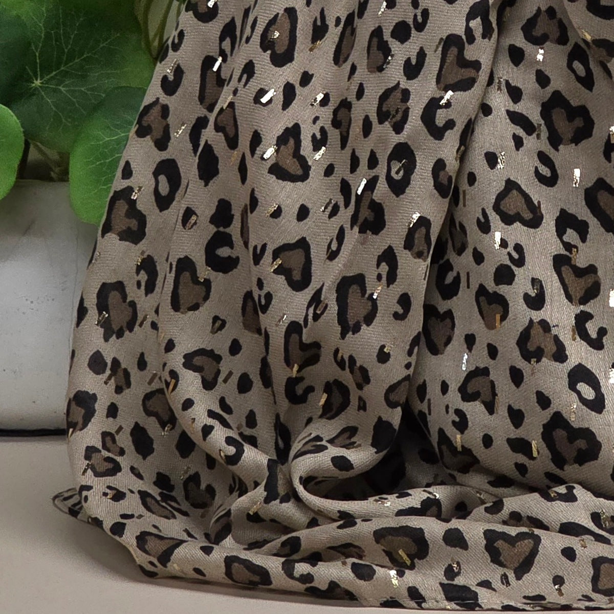 A draped leopard print scarf with a beige base, black and brown spots, and small scattered metallic accents, shown against a neutral surface with a blurred green plant in the background.