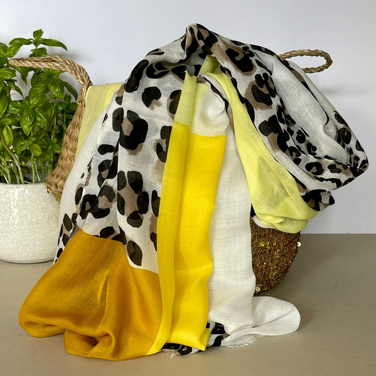 A white, black and beige leopard print scarf draped over a woven basket, featuring block colours of mustard, yellow and light yellow.  Set against a neutral background with a potted plant.
