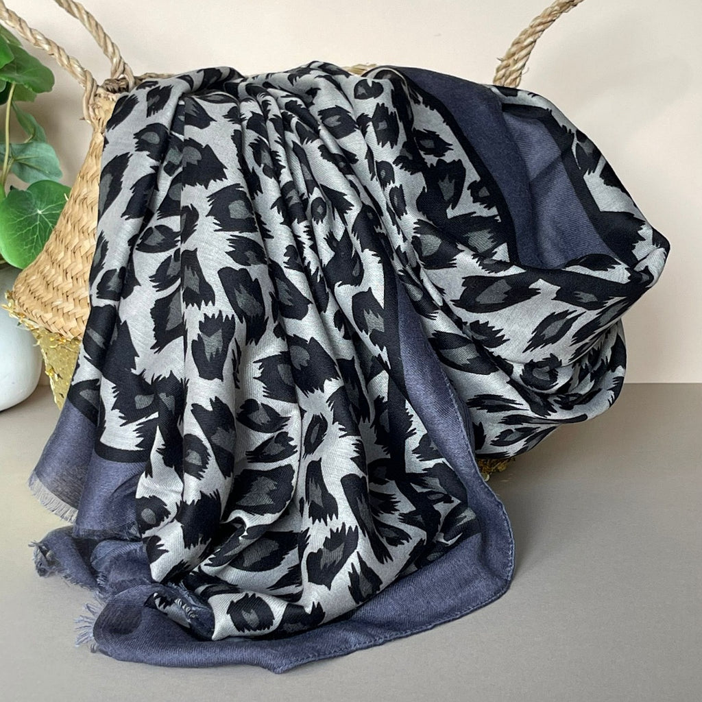 A draped scarf with a grey, black, and dark grey leopard print pattern, featuring a contrasting black and dark grey border, styled in a basket.