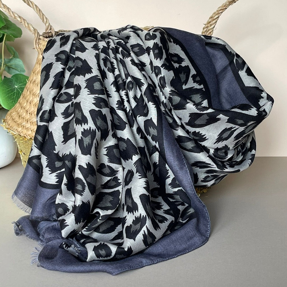 A draped scarf with a grey, black, and dark grey leopard print pattern, featuring a contrasting black and dark grey border, styled in a basket.