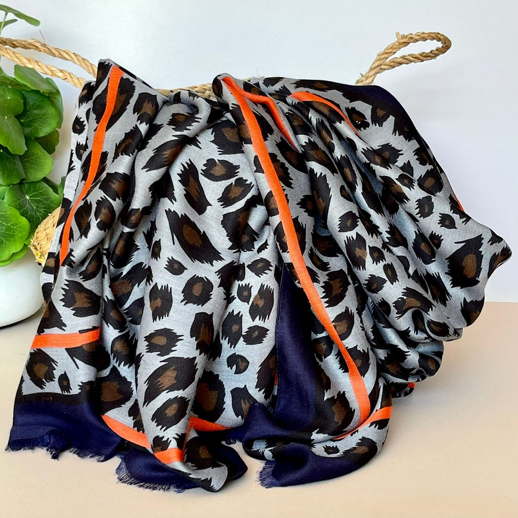 A draped scarf with a grey, black, and brown leopard print pattern, featuring a contrasting orange and navy border, styled in a basket.