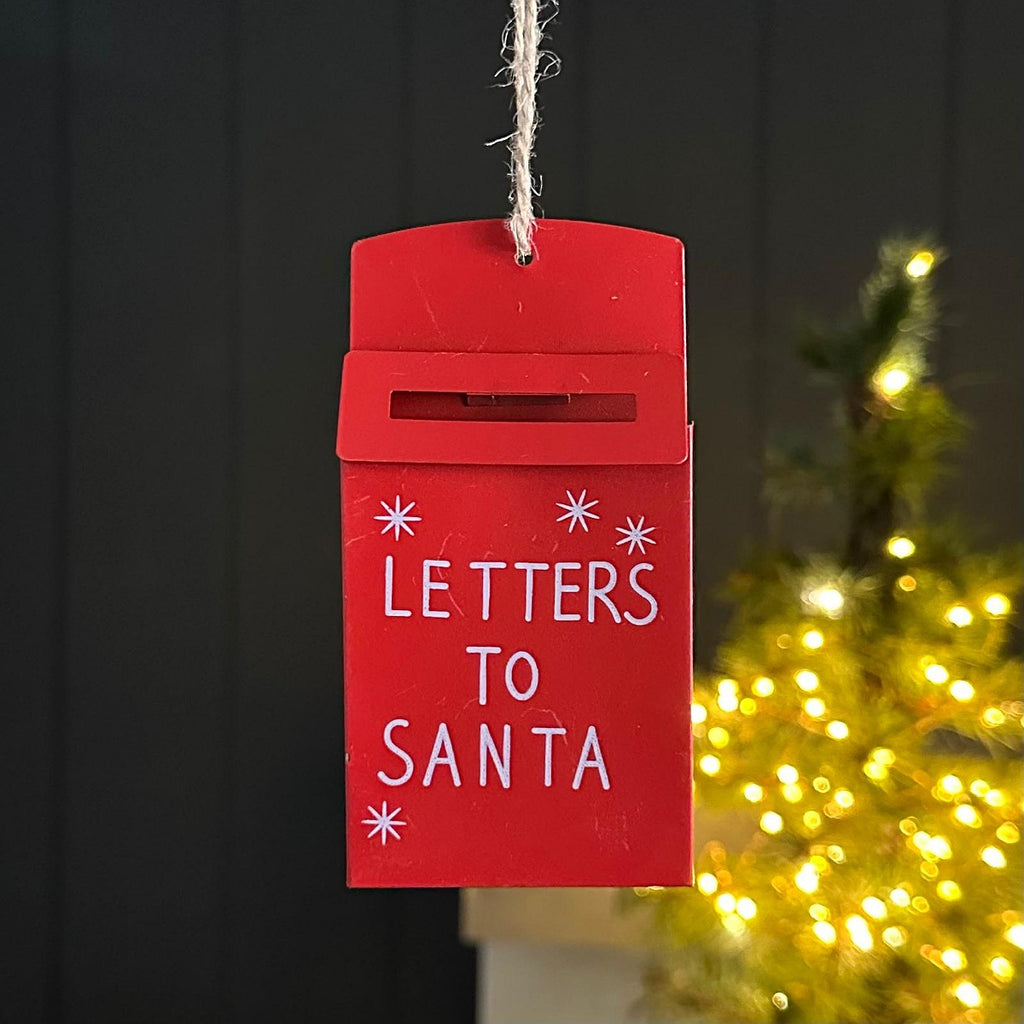 A red metal Christmas hanging ornament shaped like a mailbox with the words 'LETTERS TO SANTA' in white, hanging by a string against a dark panelled wall with a blurred Christmas tree in the background.