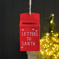 A red metal Christmas hanging ornament shaped like a mailbox with the words 'LETTERS TO SANTA' in white, hanging by a string against a dark panelled wall with a blurred Christmas tree in the background.