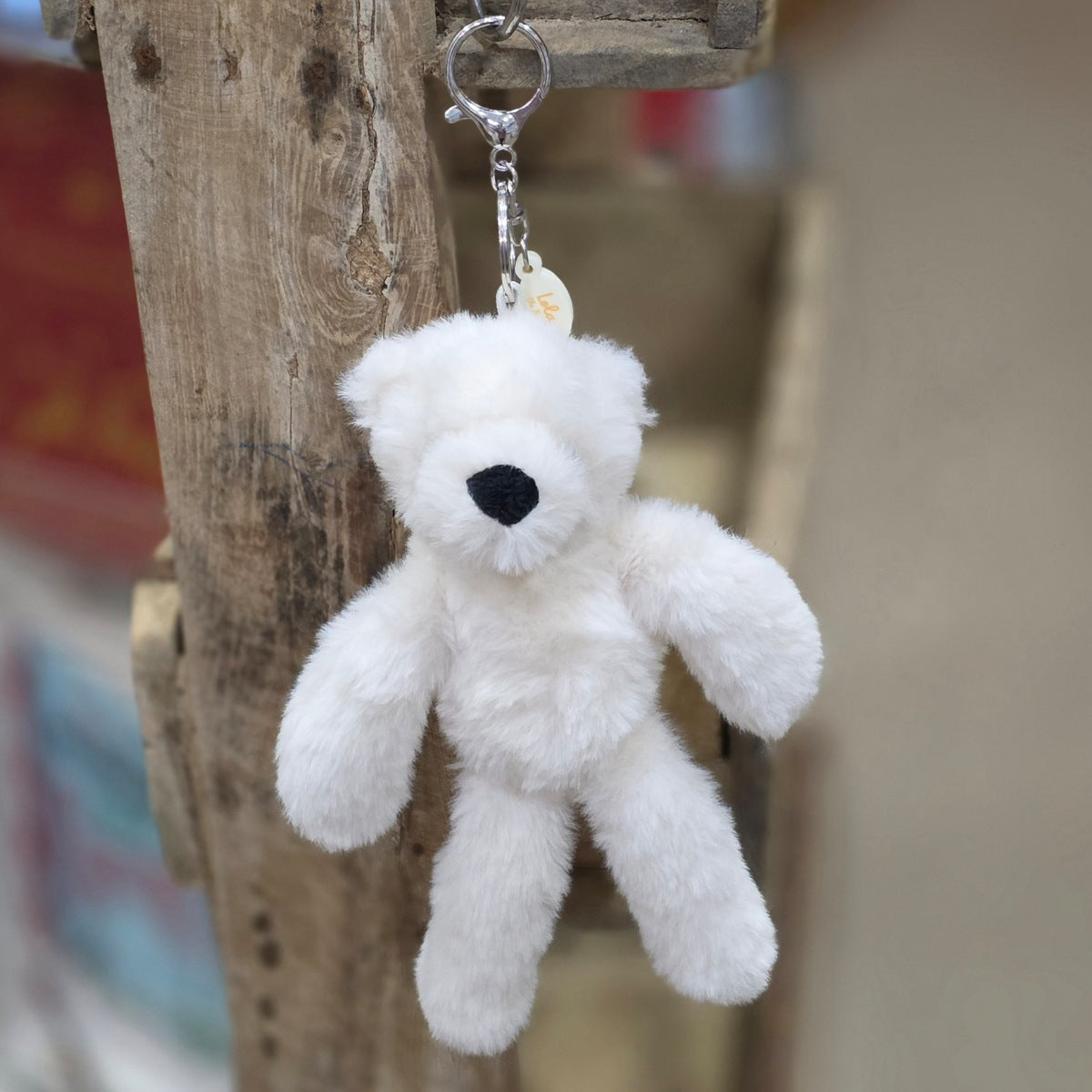 A cream plush teddy bear keychain hanging from a rustic wooden post. The keychain features a soft, fuzzy cream bear with black nose, attached to a silver metal key ring and a small round tag.
