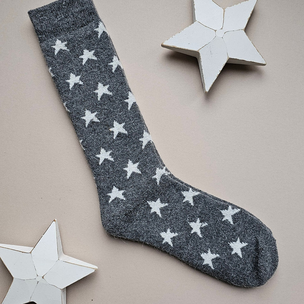 A single dark grey sock with an all-over pattern of white stars, laid flat on a light beige background with decorative white star cutouts.