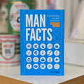 A blue hardcover book titled "MAN FACTS" by Dan Bridges stands upright on a white surface with blurred cans in the background.