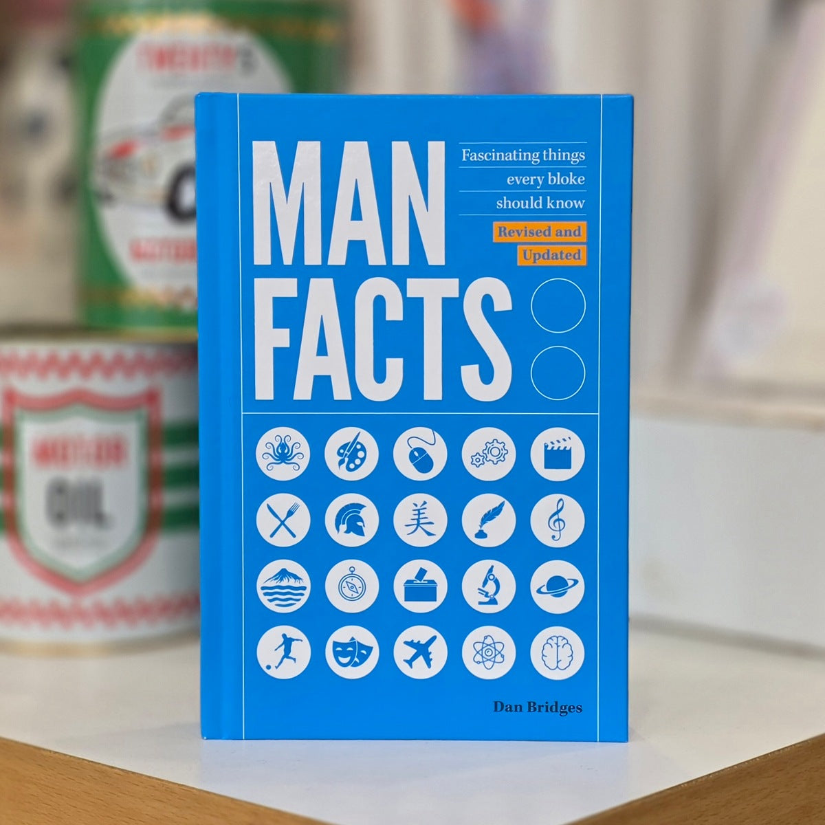 A blue hardcover book titled "MAN FACTS" by Dan Bridges stands upright on a white surface with blurred cans in the background.