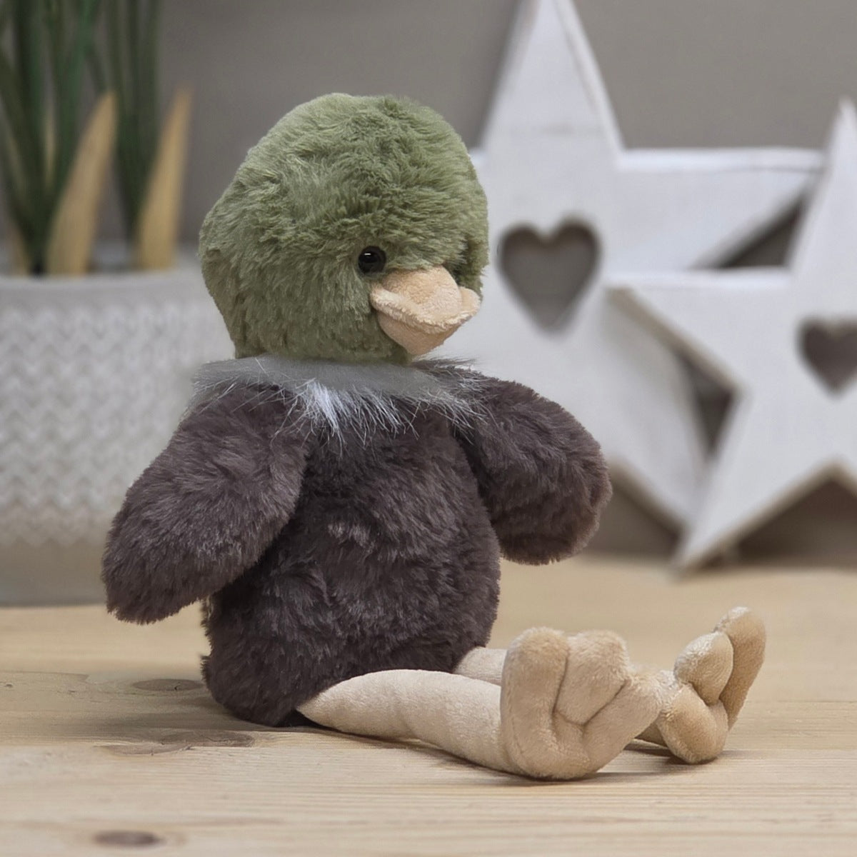 A brown soft fuzzy plush mallard with black eyes, olive green round head, white fluffy collar, beige long legs and beak.  On a wooden surface with a blurred star-shaped object in the background