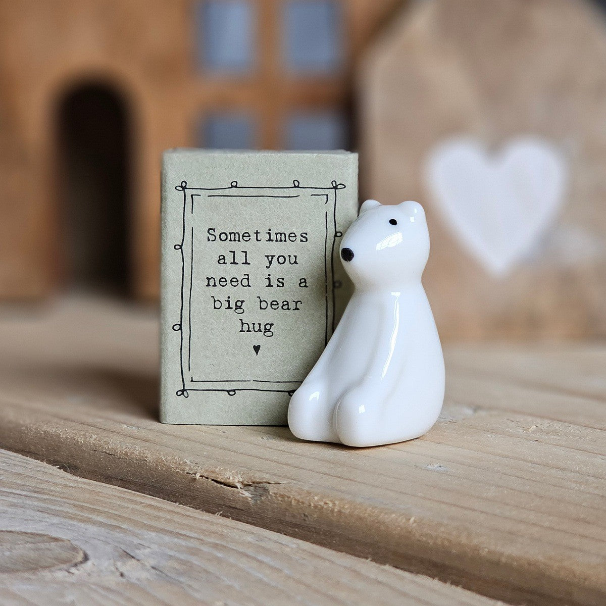A small white ceramic bear figurine stands beside its light grey packaging box on a wooden surface. The box is printed with the words 'Sometimes all you need is a big bear hug' and a black heart.