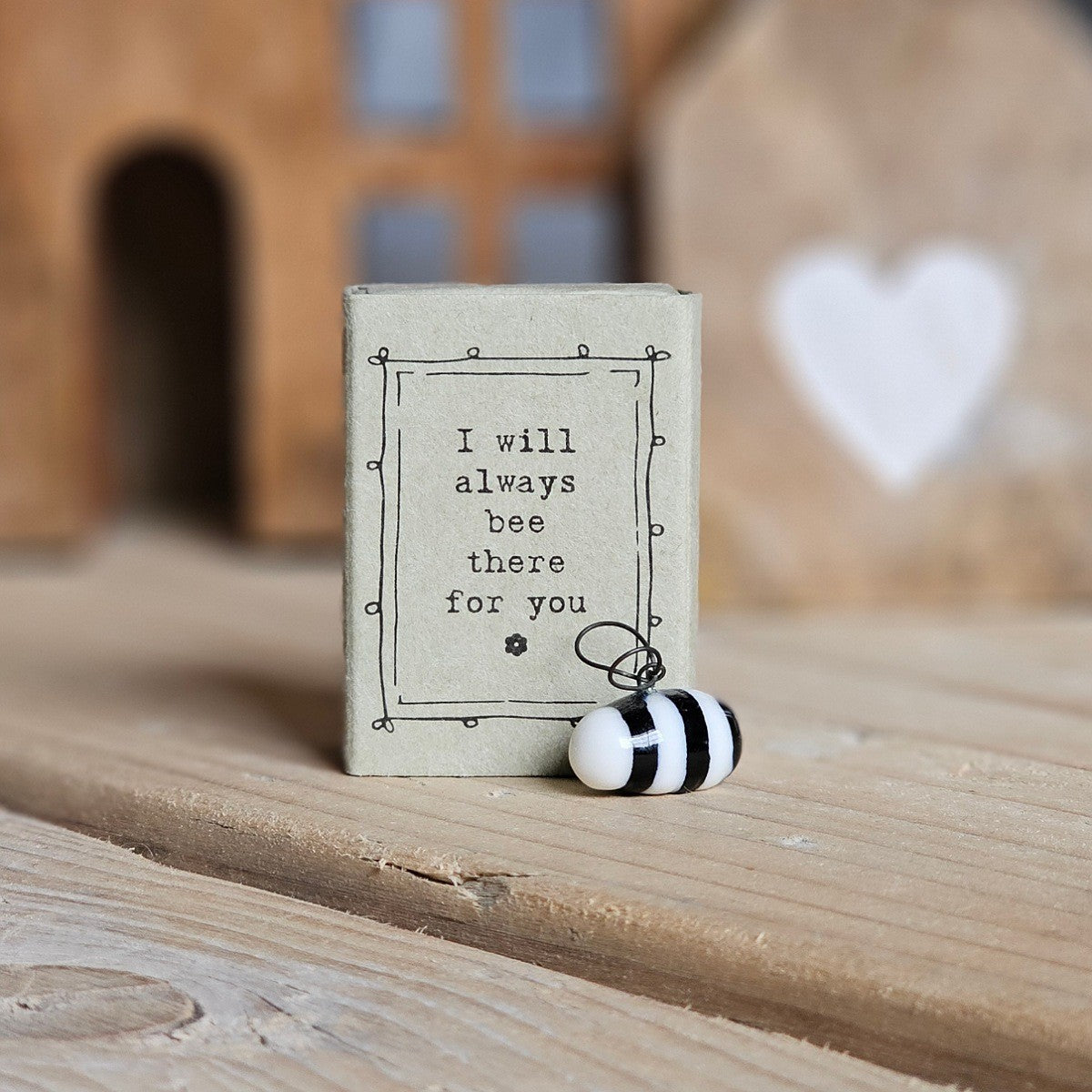 A small white ceramic bee figurine stands beside its light grey packaging box on a wooden surface. The box is printed with the words 'I will always bee there for you' and a black flower.