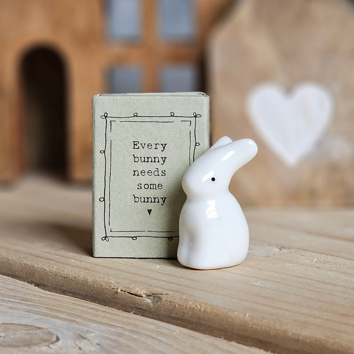 A small white ceramic bunny figurine stands beside its light grey packaging box on a wooden surface. The box is printed with the words 'Every bunny needs some bunny' and a black heart.