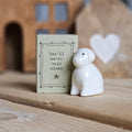 A small white ceramic dog figurine stands beside its light grey packaging box on a wooden surface. The box is printed with the words 'You'll never walk alone' and a black flower.