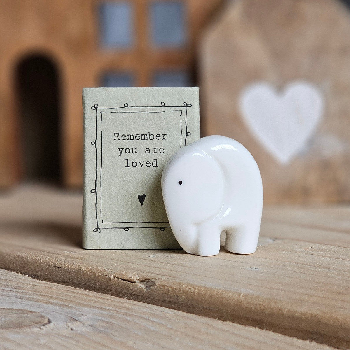 A small white ceramic elephant figurine stands beside its light grey packaging box on a wooden surface. The box is printed with the words 'Remember you are loved' and a black heart.