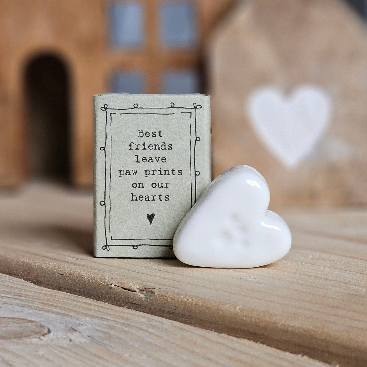 A small white ceramic heart with paw print figurine stands beside its light grey packaging box on a wooden surface. The box is printed with the words 'Best friends leave paw prints on our hearts' and a black heart.